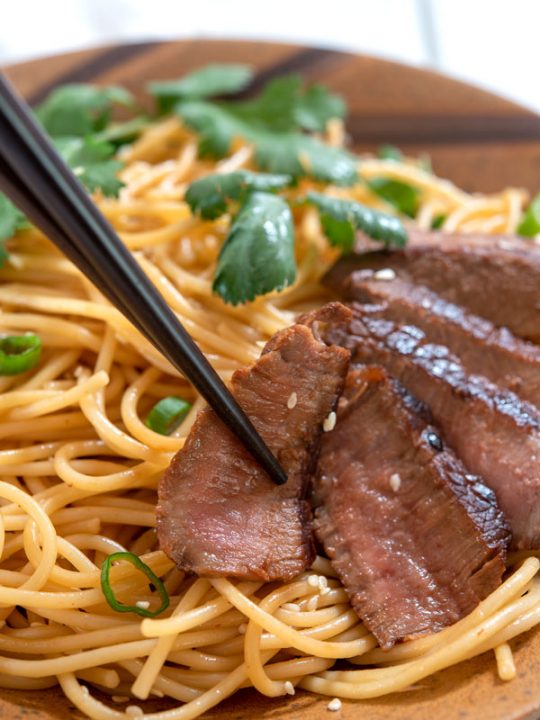 Chopsticks picking up slice of beef steak with sesame noodles