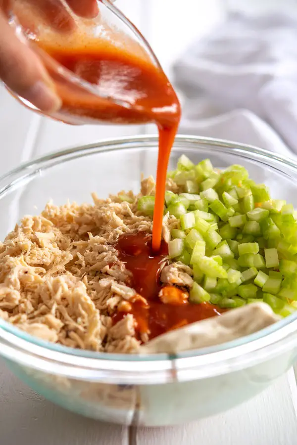 Glass bowl of buffalo chicken roll ups filling: chopped chicken, celery and cheese being drizzled with hot sauce