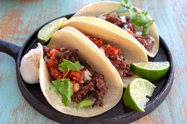 3 barbacoa tacos on a black plate topped with salsa, cilantro and onion