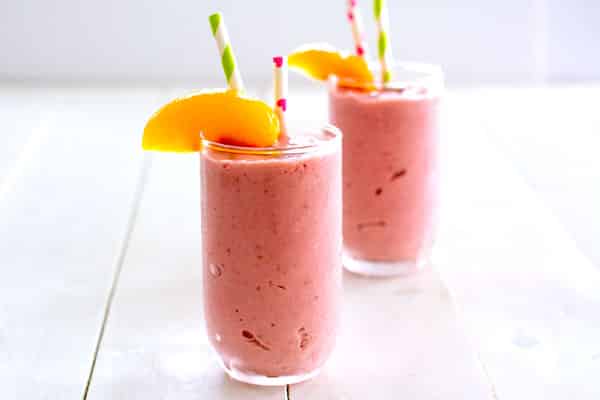 Two clear glasses filled with pink Raspberry Peach Cottage Cheese Smoothie garnished with straws and a peach slice