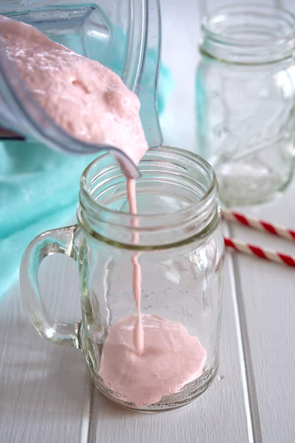 Strawberry milkshake without ice cream being poured from blender to mason jar cup