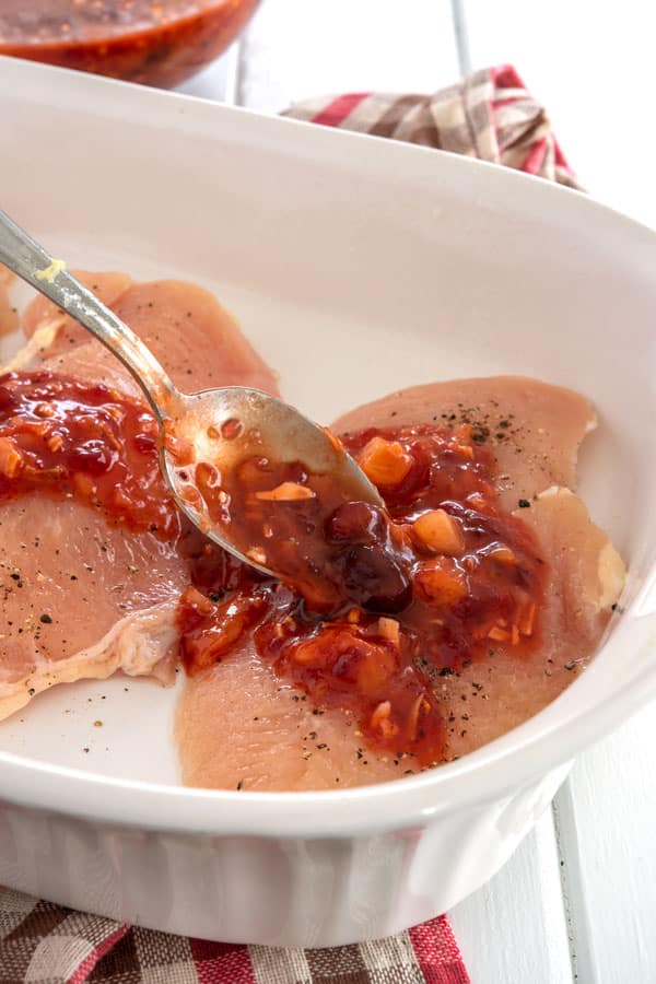 Baking dish with raw chicken breasts being spooned with cranberry sauce before baking