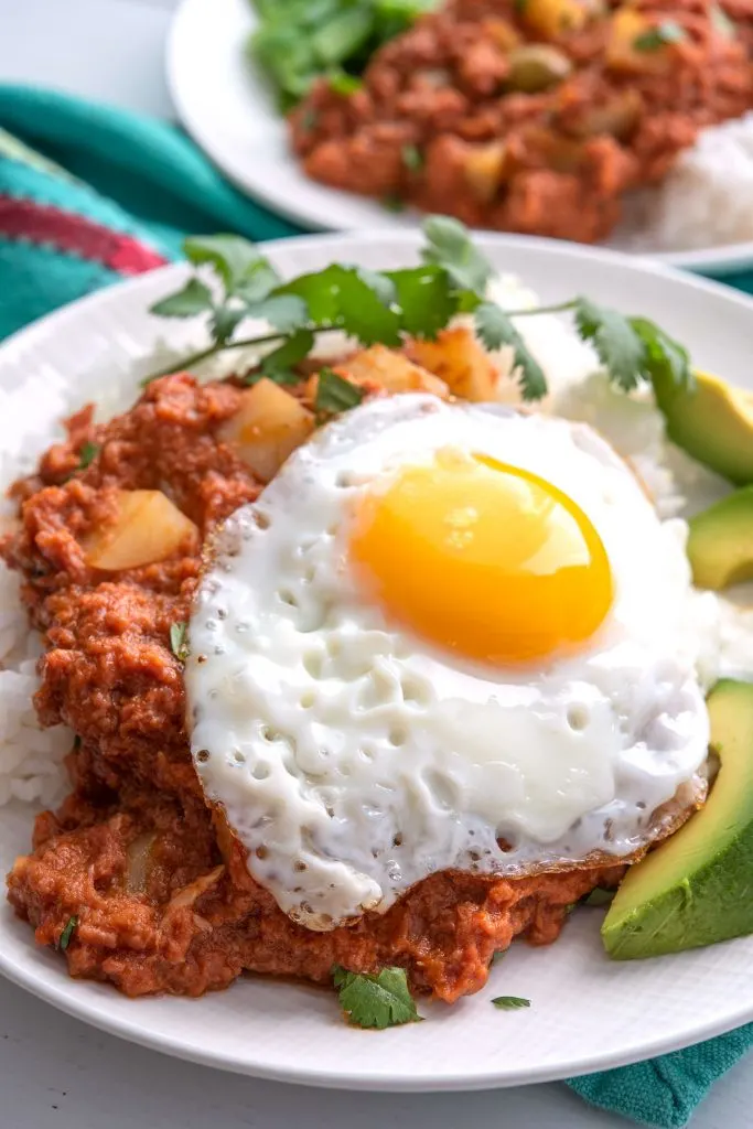 Plate of corned beef over rice with fried egg on top and avocado on the side